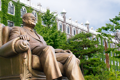 김인곤 선생의 동상이 학교 전경을 배경으로 클로즈업 되어있다.